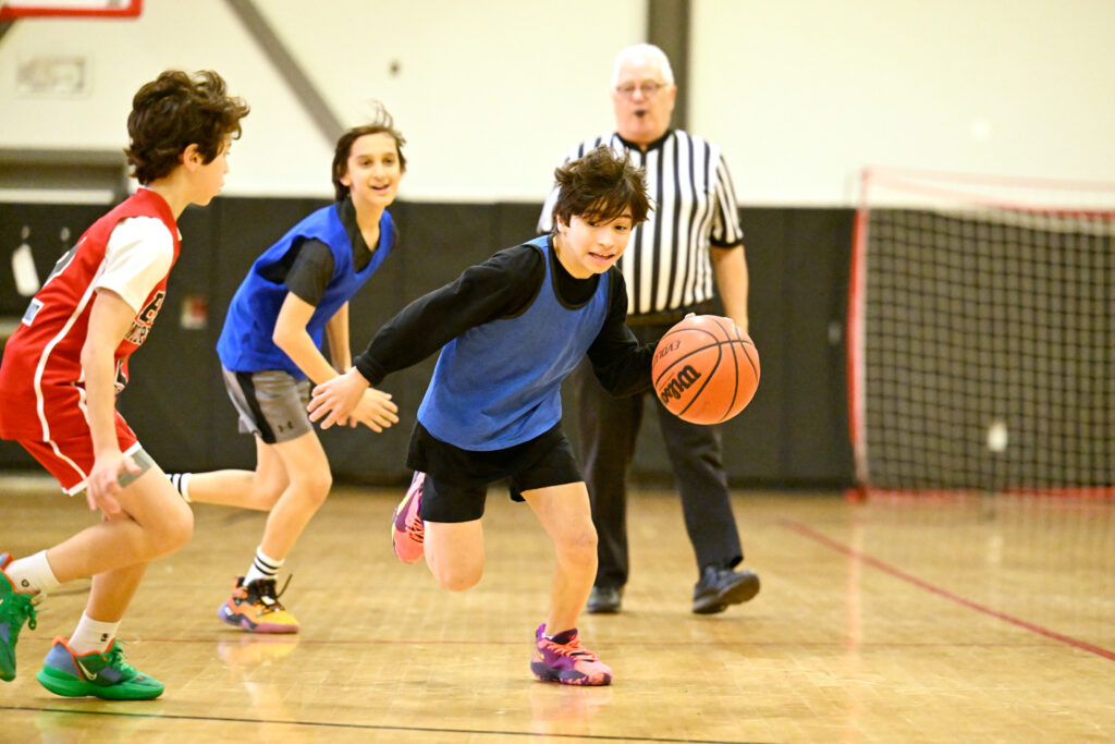 two middle school boys playing basketball and running with an opposing player encroaching and a referee in the background