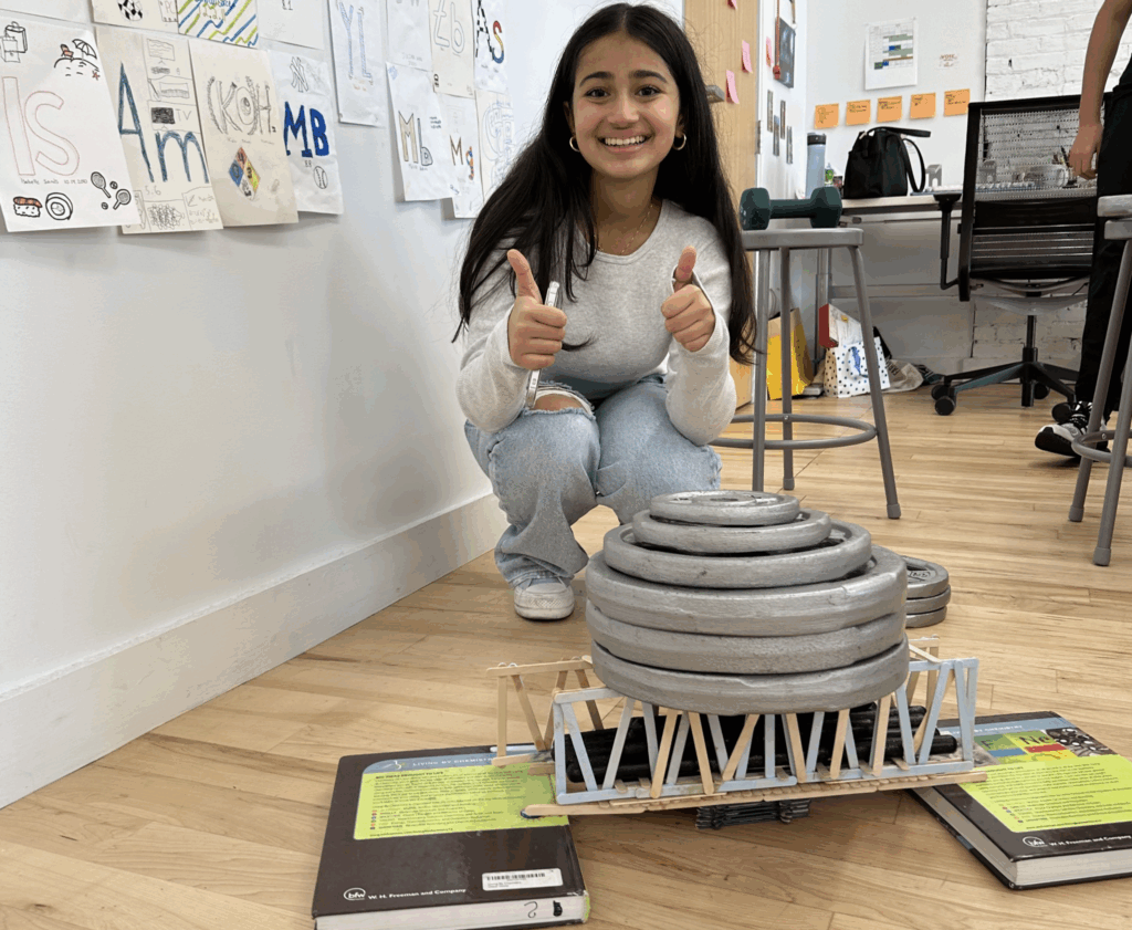 a female student gives two thumbs up after testing the bearing weight her bridge could hold in engineering club. she built it with popsicle sticks and stacked six metal weights on top