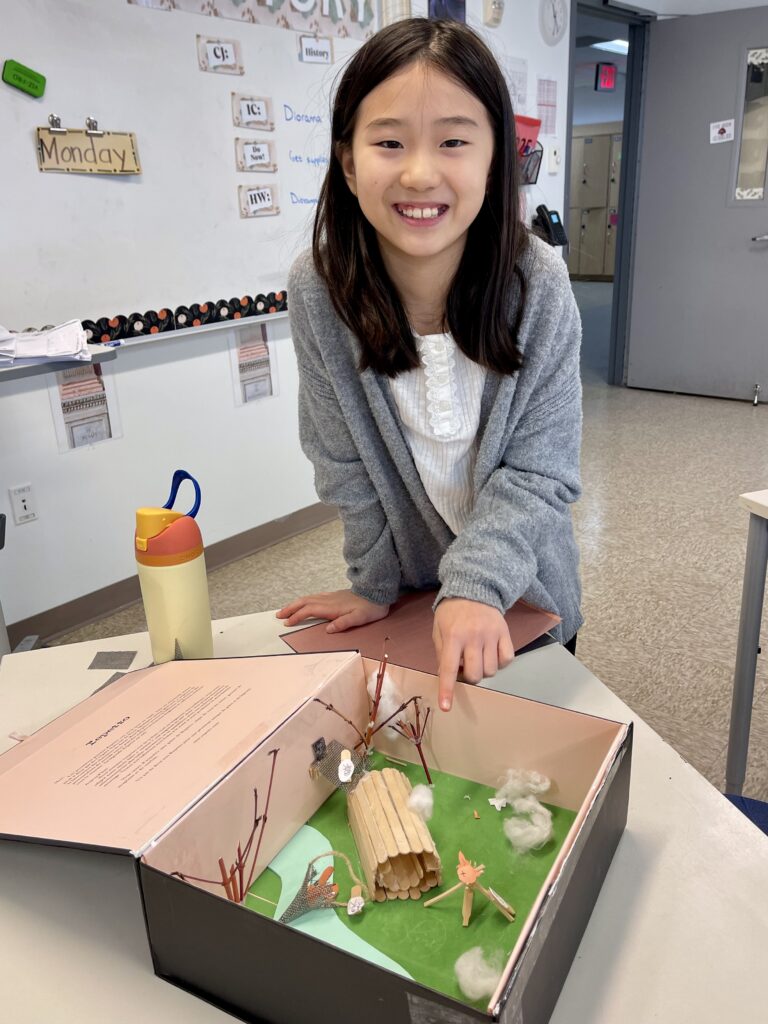 female student pointing to her diorama project in a shoe box with typed text on the top and then the native american society landscape inside the box.