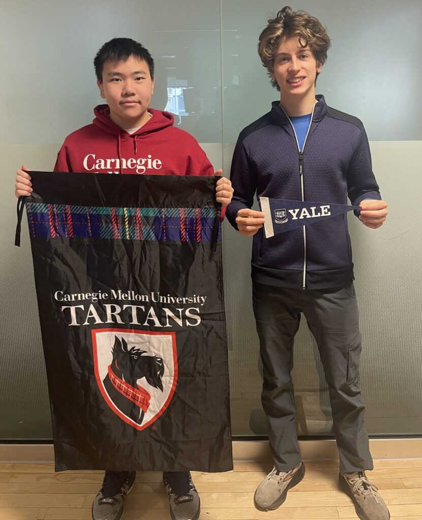 two male senior students hold up college flags to show where they are enrolled -- one says Carnegie Mellon University and the other one says Yale.