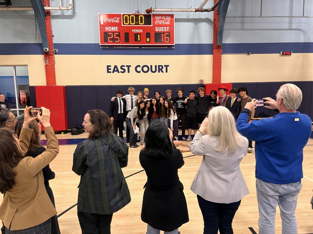 juniors and seniors smile in a group standing on the gym floor at Chelsea Piers while their parents are in front of them taking pictures