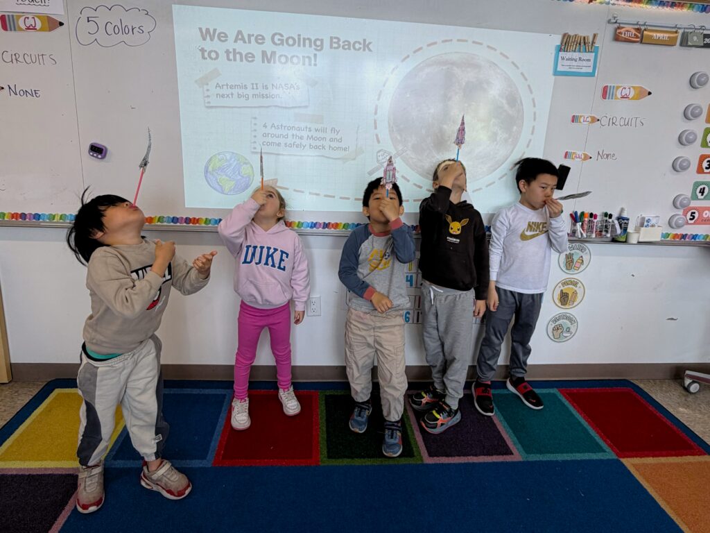 five students stand in front of a white board that says "We are going back to the moon" and they are holding their straw rockets.