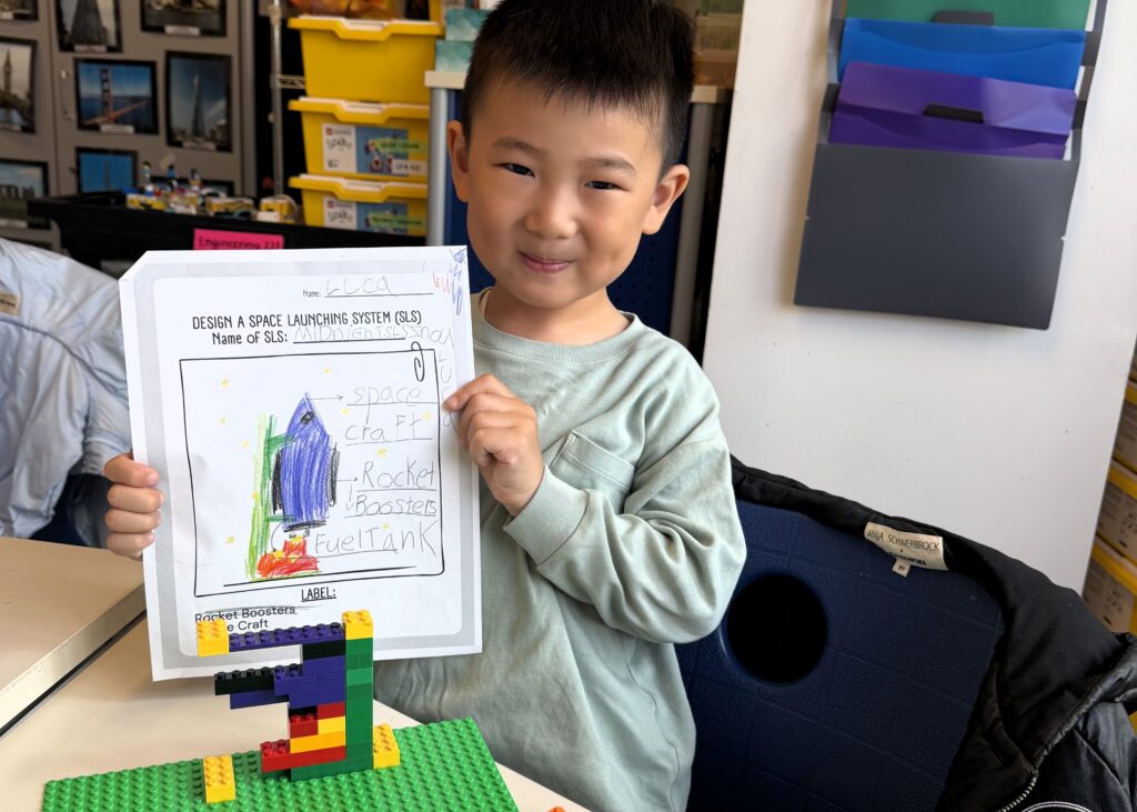 a boy student holds up a design of a space launching system with the rocket and parts labeled in front of a LEGO model.